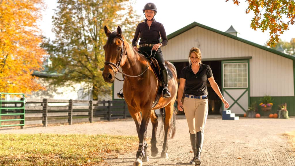 Photo of a smiling person riding a horse next to their personal trainer at Still Water Farm on a sunny autumn day