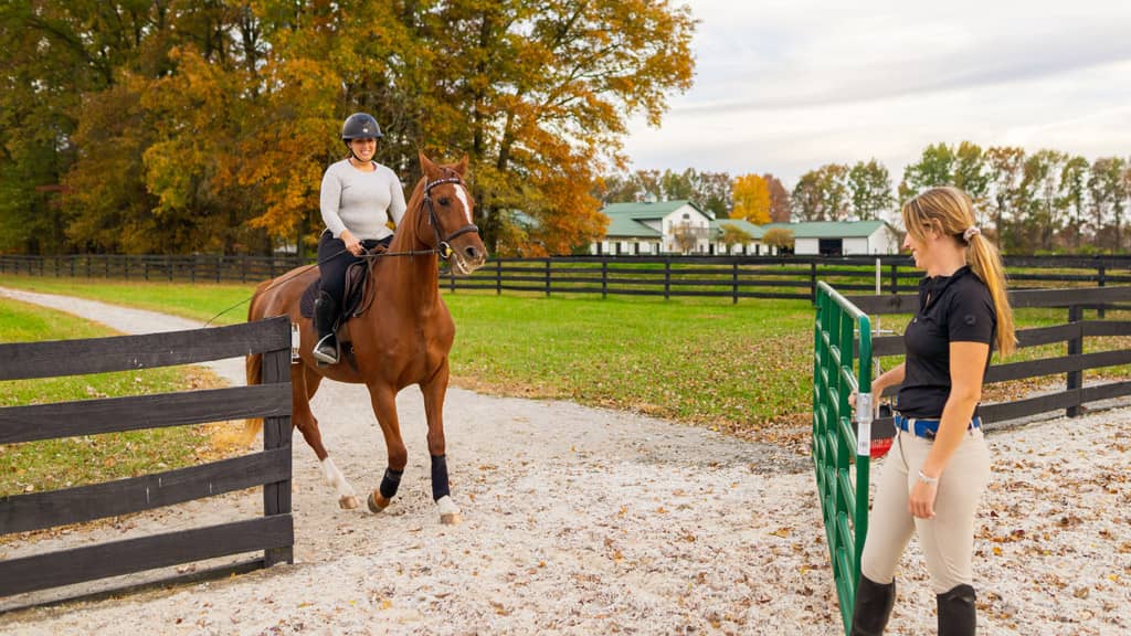 Smiling woman riding a horse into an outdoor riding arena with a gate held open by a trainer at Still Water Farm